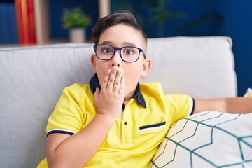 Canvas Print - Young hispanic kid sitting on the sofa at home covering mouth with hand, shocked and afraid for mistake. surprised expression