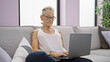 © Krakenimages.com - Focused elderly woman with grey hair and glasses, using laptop with concentration while sitting on sofa indoors at home, embodying mature technology use in the living room