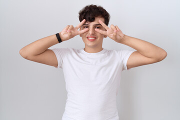 Canvas Print - Young non binary man wearing casual white t shirt doing peace symbol with fingers over face, smiling cheerful showing victory