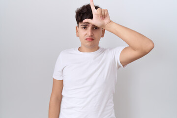 Young non binary man wearing casual white t shirt making fun of people with fingers on forehead doing loser gesture mocking and insulting.