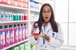 Young hispanic woman working at pharmacy drugstore holding syrup in shock face, looking skeptical and sarcastic, surprised with open mouth