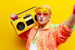 © Богдан Маліцький - old crazy grandmother in fashionable sportswear listens loudly to music on a tape recorder and takes a selfie on yellow isolated background, elderly funny pensioner in glasses holds a boombox and