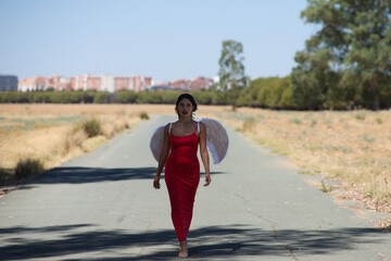 Beautiful young Latin woman in red silk dress and white wings walks along a tarmac road in the countryside outside the big city. The woman makes different poses and expressions.