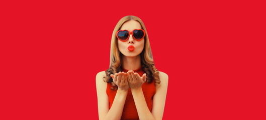 Wall Mural - Portrait of beautiful young woman blowing her lips with red lipstick sends sweet air kiss wearing heart shaped sunglasses on studio background