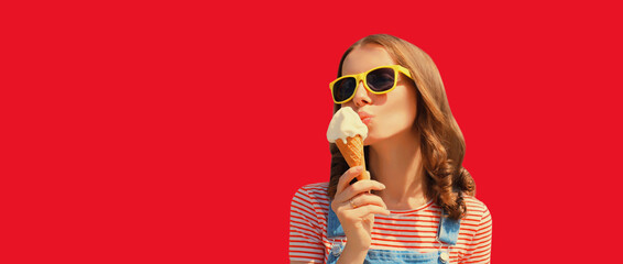 Wall Mural - Summer portrait of happy young woman eating ice cream wearing sunglasses on red studio background