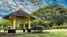 Gazebo For Relaxing In The Tropical Garden. Conical Roof, Columns, Wooden Benches. Flowering Bushes On A Green Lawn. Green Trees Against A Blue Sky And Clouds. Malaysia. Borneo. Kota Kinabalu.