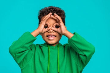 Wall Mural - Photo of cheerful carefree cute boy dressed trendy clothes showing gesture looking through hole isolated on cyan color background