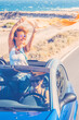 © simona - Happy women traveler enjoy road trip with a convertible car. One woman outside the roof have fun. Scenic desert destination. Car driving. Excited couple of friends in a vehicle on travel. Adventure