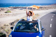 © simona - Happy women traveler enjoy road trip with a convertible car. One woman outside the roof have fun. Scenic desert destination. Car driving. Excited couple of friends in a vehicle on travel. Adventure