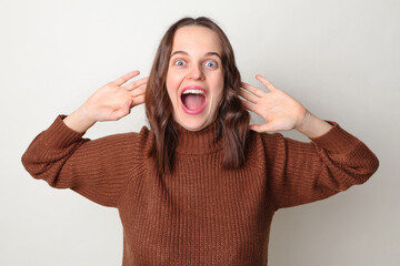 Wall Mural - Cheerful funny surprised brown haired adult woman wearing brown sweater posing isolated over gray background raised her arms screaming with amazement exclaiming