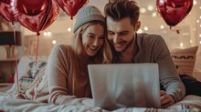 Happy loving couple shopping online while lying in bed and using laptop with heart shape balloons