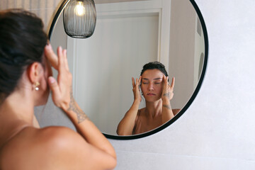 Young Middle Eastern woman wearing a silk nightdress, looking at the mirror and doing face sculpting massage. Morning beauty routine concept. Close up, copy space, background.