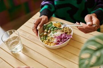 Wall Mural - cropped photo of african american woman holding fork with corn while eating vegan bowl in cafe