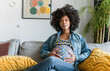 © Victor - Calm young pregnant woman sitting on sofa and embracing her belly. Portrait of pregnant woman resting at her cozy home. Concept of motherhood and expecting baby.