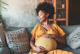Fototapeta  - Calm young pregnant woman sitting on sofa and embracing her belly. Portrait of pregnant woman resting at her cozy home. Concept of motherhood and expecting baby. 