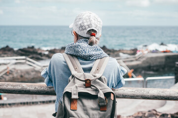 Sticker - Rear view of senior traveler woman with cap looking at horizon over sea, elderly caucasian lady carrying backpack enjoying nature and vacation