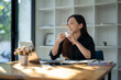 © Wasana - Freelance Asian woman enjoying morning coffee during break from work with sunlight shining through the glass on her table.
