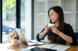 © Wasana - Freelance Asian woman enjoying morning coffee during break from work with sunlight shining through the glass on her table.