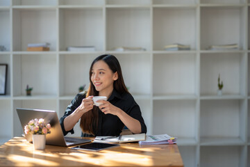 Freelance Asian woman enjoying morning coffee during break from work with sunlight shining through the glass on her table.