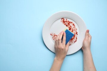 Sticker - Woman washing dirty plate with sponge on light blue background, top view. Space for text