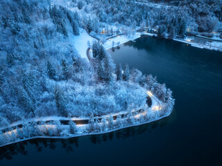 Wall Mural - Aerial view of road, snowy forest, illumination, lake, street lights at winter night. Top drone view of alpine countryside, road through the snowy pine trees at dusk. Bled lake, Slovenia. Travel