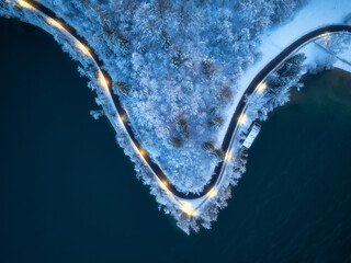 Wall Mural - Aerial view of road, snowy forest, illumination, lake, street lights at winter night. Top drone view of alpine countryside, road through the snowy pine trees at dusk. Bled lake, Slovenia. Travel