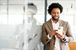 A business-savvy African-American man engages with his smartphone, his reflection mirroring the modern multitasking professional in a glass office setting. Mobile technology in corporate world concept