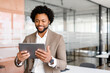 © Vadim Pastuh - A confident young businessman in a tan suit smiles while holding a tablet, standing in a modern office environment. Concept of themes of business, technology, and entrepreneurship.