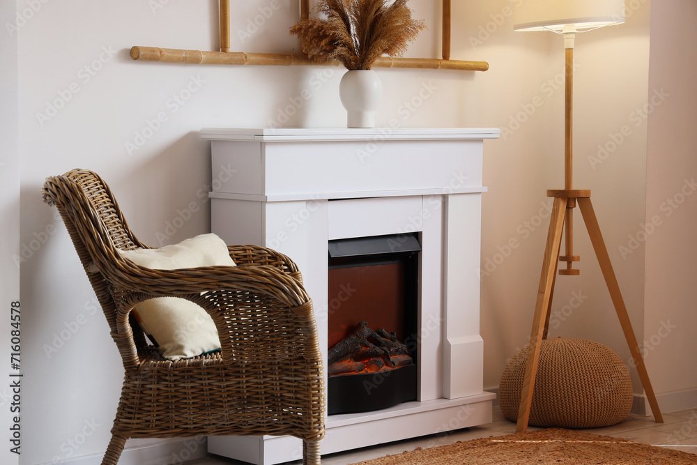 Stylish fireplace, wicker armchair and lamp near light wall