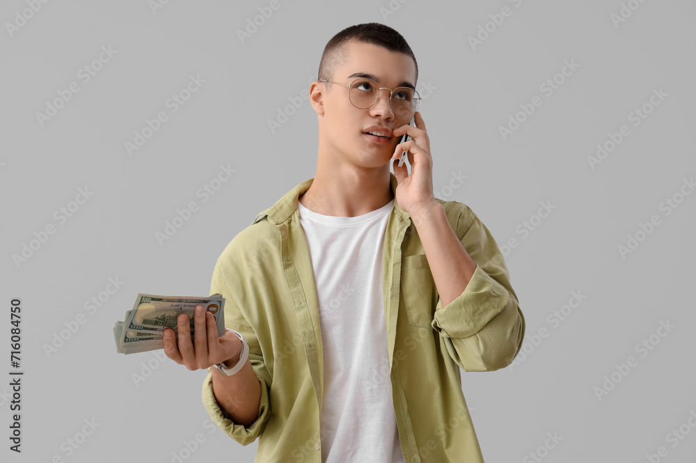 Young man with dollar banknotes talking by mobile phone on light background