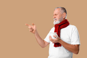 Wall Mural - Old man with sore throat holding cup of tea and pointing at something on beige background