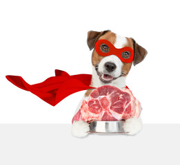 Wall Mural - Funny jack russell terrier puppy wearing superhero costume holds bowl of raw meat and looks above empty white banner. isolated on white background