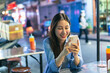 Young Asian woman traveler tourist using a smart phone while eating Thai street food in China town night market in Bangkok in Thailand - people traveling enjoying food culture concept