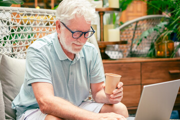 Wall Mural - Portrait of smiling relaxed bearded senior man using laptop computer sitting indoor on armchair. Old generation people and new technologies