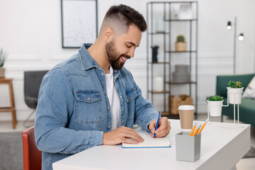 Wall Mural - Young man writing in notebook at white table indoors