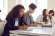 © Studio Romantic - Side view of a group of diverse young high school students boys and girls sitting at the desk in a row and writing test during a college lesson in the classroom. Education and knowledge concept.