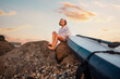 © _KUBE_ - Summer activity vacations. Pre-school girl sitting with sup board at big beach rocks. Bottom view. Sunset sky at the background