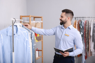 Wall Mural - Dry-cleaning service. Happy worker with notebook choosing clothes from rack indoors