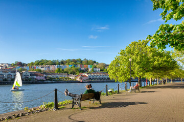 Wall Mural - Bristol, UK - A beautiful spring evening at Bristol Docks, young couple sitting on a bench, colourful houses, fresh green trees, blue sky, sail boat on the water.