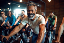 Sporty Mature African American Man In Workout Attire Cycles On A Stationary Bike In A Diverse Gym Class With Others.