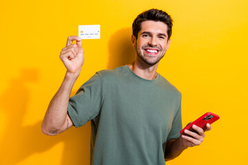 Poster - Photo of optimistic cheerful man dressed khaki t-shirt hold smartphone demonstrate credit card isolated on yellow color background