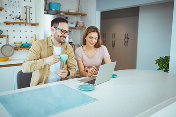 Pleasant family couple sitting at big wooden table in modern kitchen, looking at laptop screen. Happy young mixed race married spouse web surfing, making purchases online or booking flight tickets.
