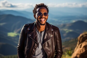 Wall Mural - Portrait of a cheerful afro-american man in his 30s sporting a classic leather jacket against a panoramic mountain vista. AI Generation