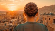  The Back Of A Woman's Head As She Stands In Front Of A View Of A City With Buildings And A Mountain In The Distance With A Sunset In The Background.