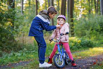 School kid boy, brother put on little preschool sister girl bike helmet on head. Brother teaching happy child cycling and having fun with learning bike. Active siblings family outdoors. Kids activity