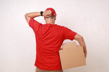 Back view of Confused Asian courier man in red holding head while carrying a cardboard box. Isolated on white