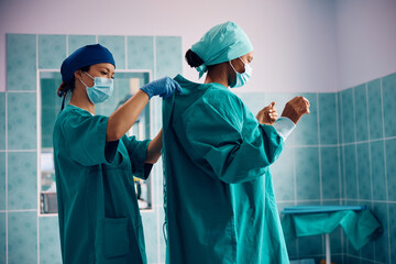 Wall Mural - Surgical nurse assisting black female doctor in getting dressed for surgery at medical clinic.