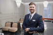 © Seventyfour - Waist up portrait of smiling young man wearing suit and looking at camera while welcoming guests in luxury hotel lobby, copy space