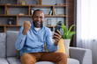 © Liubomir - Black skinned man in casual outfit sitting on couch with modern smartphone and making winner gesture in living room. Portrait of mature male rejoicing great news with satisfied facial expression.
