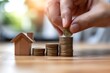 Strategic savings in action: A hand places a coin on a stack with a wooden house, illustrating the financial plan for home buying. Realize your homeownership dreams!